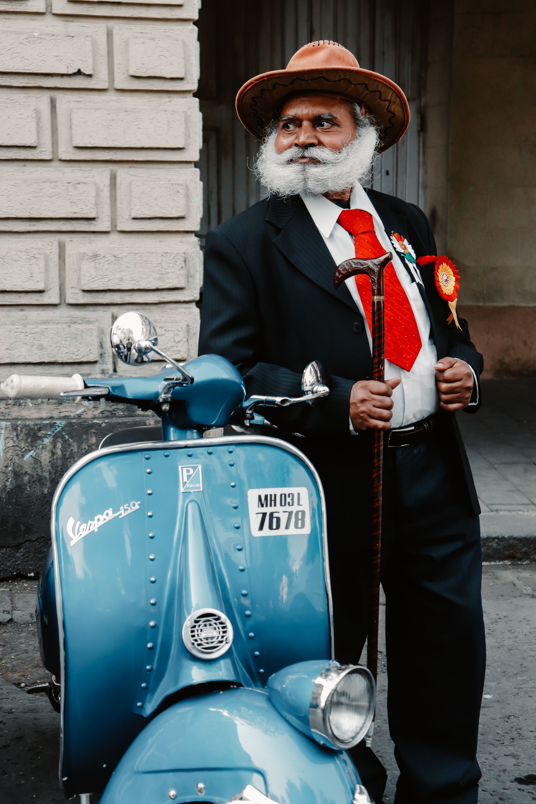 Illustrative photograph: A silver-bearded older gentleman in a smart suit and hat standing beside a vintage scooter.