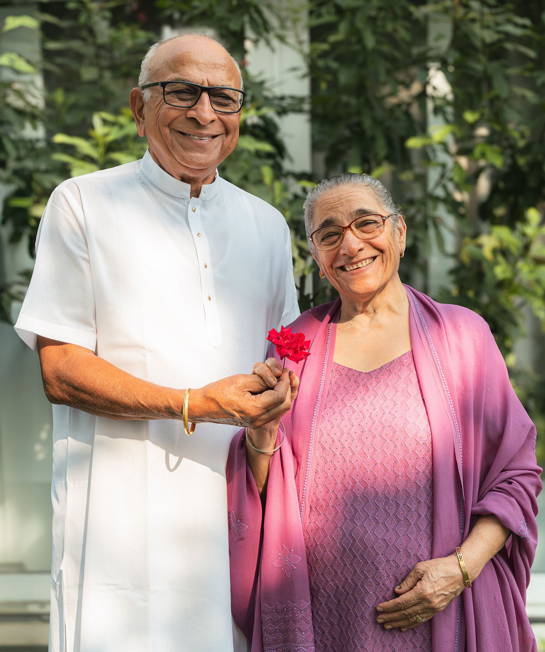 Illustrative photograph: An older Indian couple in glasses sharing a smile and holding a rose.