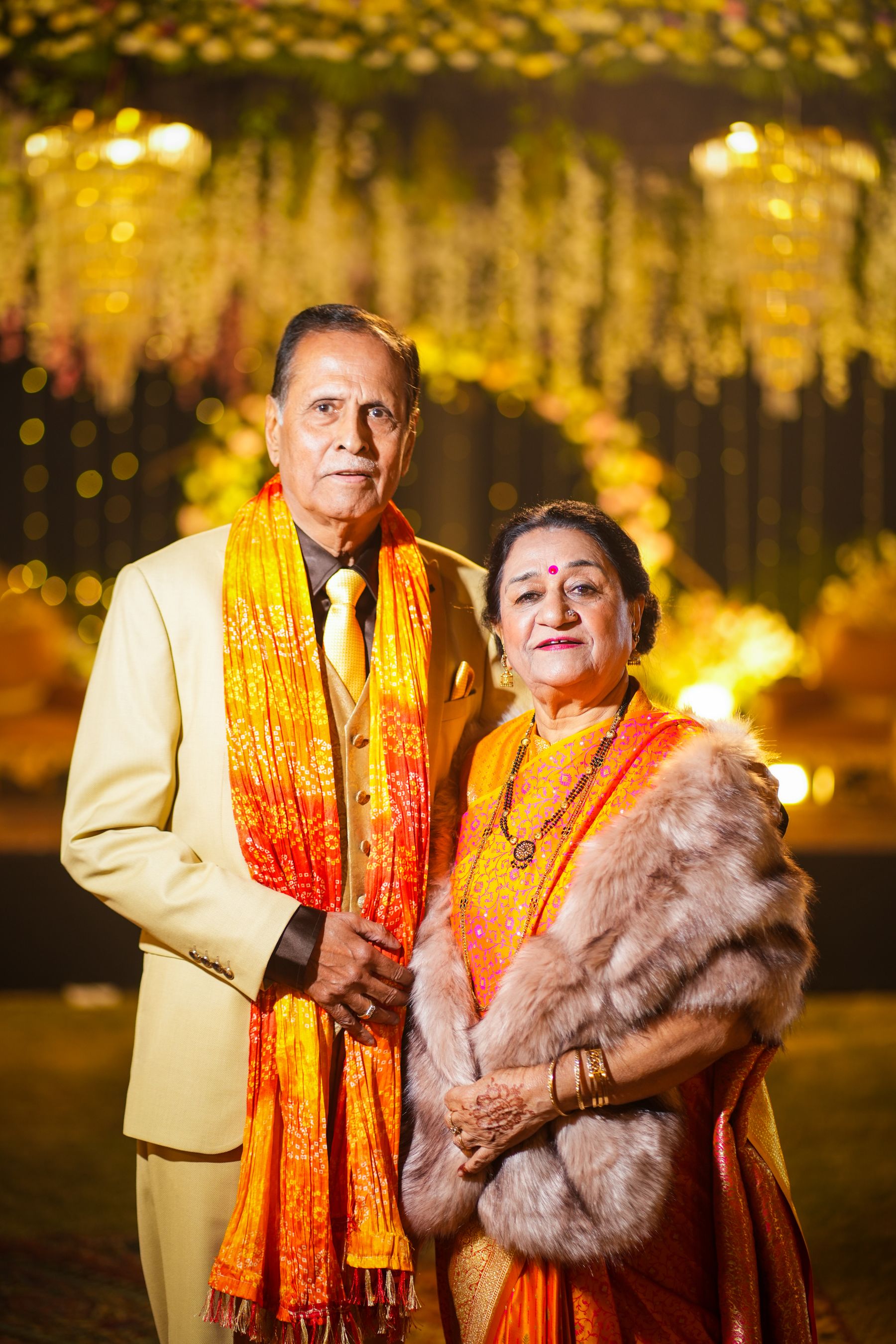 Illustrative photograph: An older couple dressed in formal Indian celebration attire, standing together in warm light.
